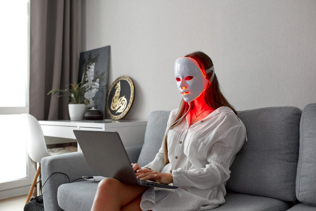 HOME a beautiful girl with an led mask on her head works at home on a laptop. home skin care concept.