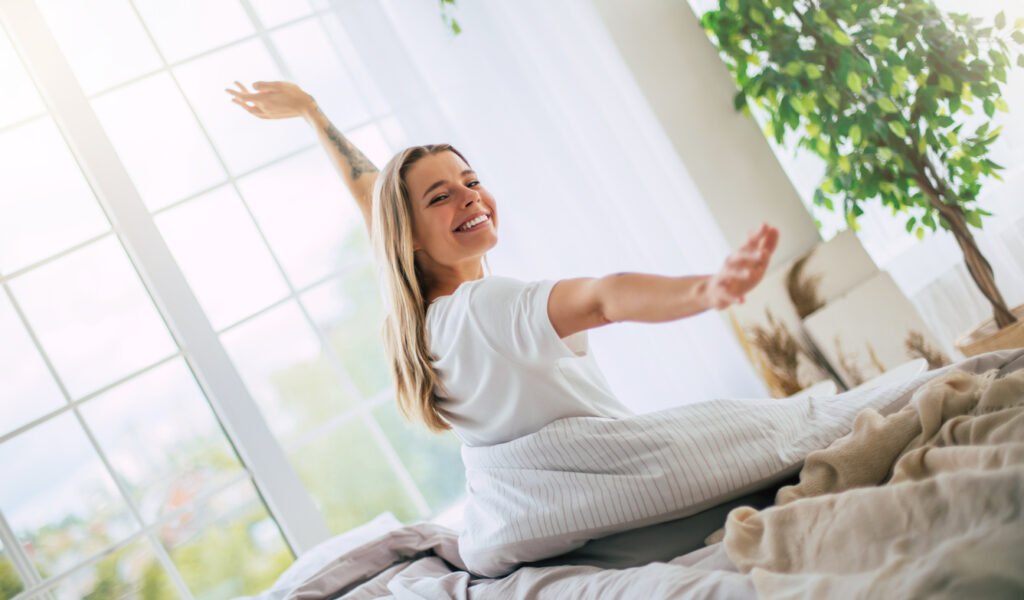 HOME cute smiling woman stretches joyfully on a bed, bathed in natural light from large windows. the bedroom is bright and cozy, with plants and soft bedding, evoking a sense of comfort and happiness